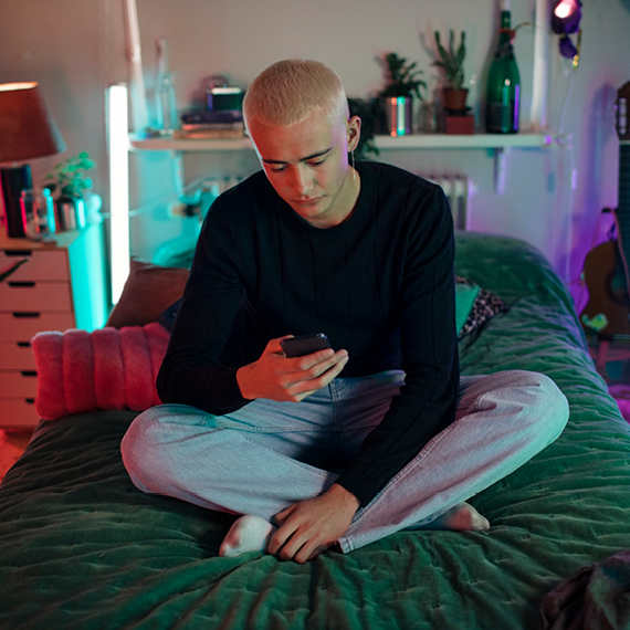 A young person, sat with cross legs on a bed, looking at a smart phone they are holding.