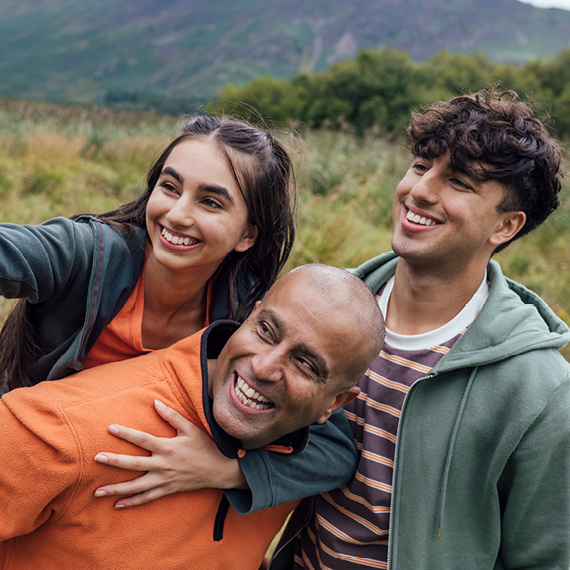 A teenage girl sits on her father's shoulders as they pose with her brother for a selfie.